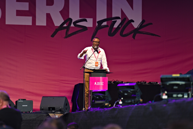 Ein Mann in einem weißen Hemd und braunen Hosen steht auf einer Bühne und hält ein Mikrofon, hinter ihm eine pinkfarbene Fahne mit der Aufschrift "Berlin as Fuck".