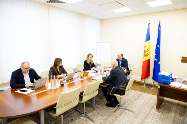 Eine Gruppe von Menschen sitzt um einen Konferenztisch mit Laptops, Flaschen, Gläsern, Papieren und Büchern, die in eine Diskussion vertieft sind, mit Flaggen, einer Whiteboard und Deckenleuchten im Hintergrund.