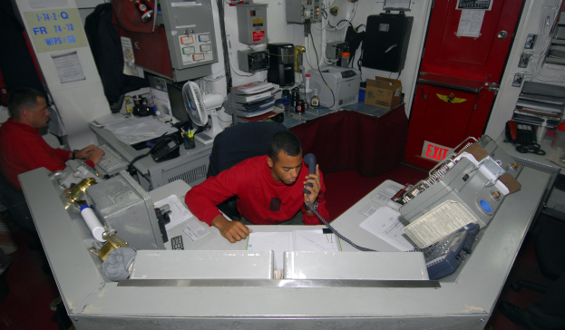 Zwei Männer sitzen an einem unordentlichen Schreibtisch und arbeiten an einem Computer, einer hält ein Telefon, mit Papieren an der Wand und einer roten Tür auf der rechten Seite.