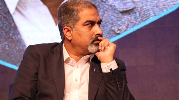 Ein Mann in Anzug sitzt auf einer Bühne und denkt nach, während er auf einen Bildschirm schaut.