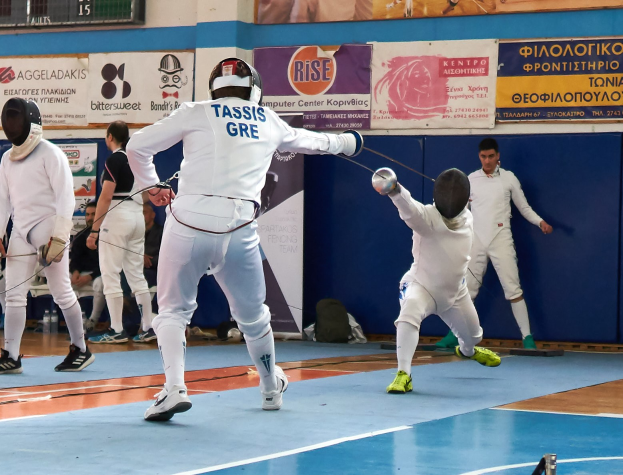 Gruppe von Fechtern in einer Turnhalle mit einer Anzeigetafel darüber, die Helme tragen und Schwerter halten, während andere im Hintergrund auf Stühlen sitzen.