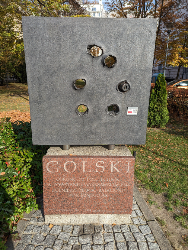 Ein Denkmal in einem Park mit Einschusslöchern, einer Pflanze auf seiner linken Seite und einem Hintergrund mit Bäumen, Gebäuden, einem Auto, einem Laternenpfahl und dem Himmel.