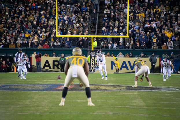 Fußballspieler mit Helmen und Uniformen auf einem Feld mit Zuschauern in Tribünen und sichtbarer Beschilderung.