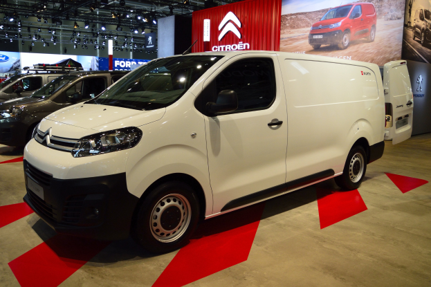 Ein weißer Citroen Berlingo Van wird auf einer Autoausstellung präsentiert, umgeben von schwarzen Schlafzimmermöbeln, während die Wände mit Brettern und Lichtern geschmückt sind.