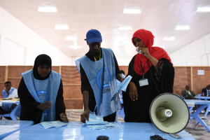 Eine Gruppe von Menschen steht um einen Tisch mit einem Megaphon und Papieren, während andere im Hintergrund eines Wahllokals mit beleuchteter Decke sitzen oder stehen.