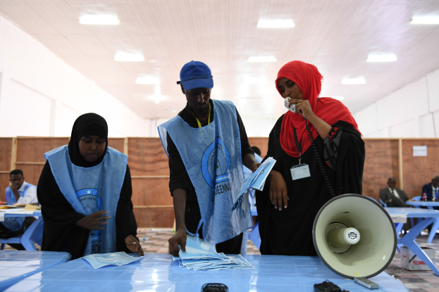 Eine Gruppe von Menschen steht um einen Tisch mit einem Megaphon und Papieren, während andere im Hintergrund eines Wahllokals mit beleuchteter Decke sitzen oder stehen.