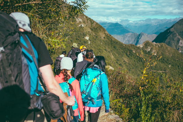 Gruppe von Menschen beim Bergwandern, mit Rucksäcken und Hüten, vor bewölktem Himmel, Bäumen und Hügeln