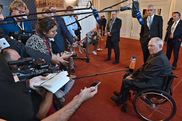 Ein Mann im Rollstuhl ist von Reportern mit Kameras, Handys und Mikrofonen mit Ständern umgeben, mit einem Banner, Scheinwerfern und einer Wand im Hintergrund.