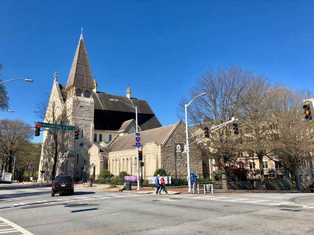 Große Kirche mit Turm an der Straßenecke, identifiziert als St. Lukes Episkopalkirche, umgeben von Gebäuden, Fahrzeugen, Fußgängern und Grün unter einem klaren blauen Himmel.