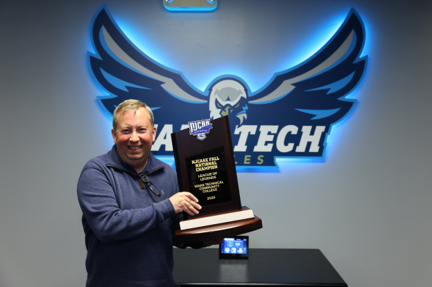 Ein Mann steht lächelnd vor einer Wand mit Logo und hält eine 'NCAA Fall National Championship'-Plakette in der Hand, im Hintergrund ist ein digitales Display auf einem Tisch zu sehen.