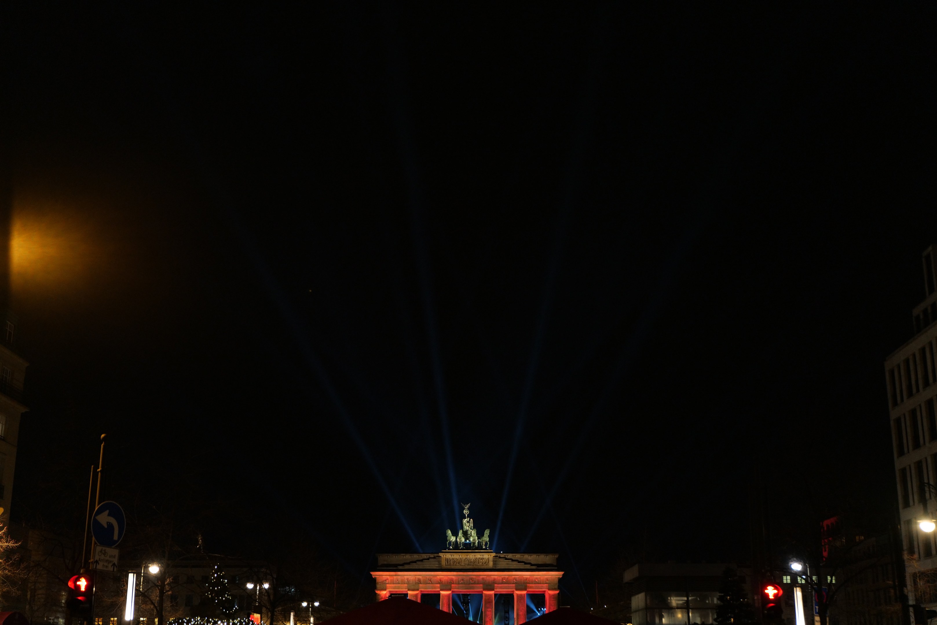 Das Brandenburger Tor nachts, beleuchtet von Straßenlaternen, mit umliegenden Gebäuden, Bäumen, Pfählen, Verkehrszeichen, Schildern und Zelten gegen einen dunklen Himmel.
