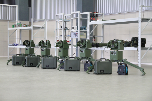 Eine Gruppe von Militärrobotern auf einem Boden mit Koffern und anderen Gegenständen auf umliegenden Regalen, vor einer Wand mit einem Geländer im Hintergrund.