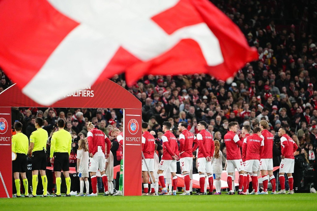 Gruppe von Menschen auf einem Fußballfeld mit einer roten und weißen Flagge im Vordergrund, einem Bogen mit dem Text "Bayern München vs. Bayern Munich Betting Tips & Preview" im Hintergrund und einer großen Menge im Stadion.