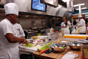 Eine Gruppe von Köchen in einer Küche beim Zubereiten von Speisen, mit einem Koch im Vordergrund in einer weißen Schürze und Mütze, der ein Messer hält und von verschiedenen Utensilien, Schüsseln, Gemüse, einem Mobiltelefon, Flaschen und anderen Gegenständen umgeben ist, mit einem Fernseher und Deckenlampen im Hintergrund.
