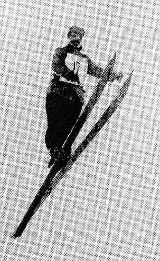 Schwarzes und weißes Foto eines Skifahrers in der Luft, der Skier hält, trägt eine Skiausrüstung mit entschlossener Miene.