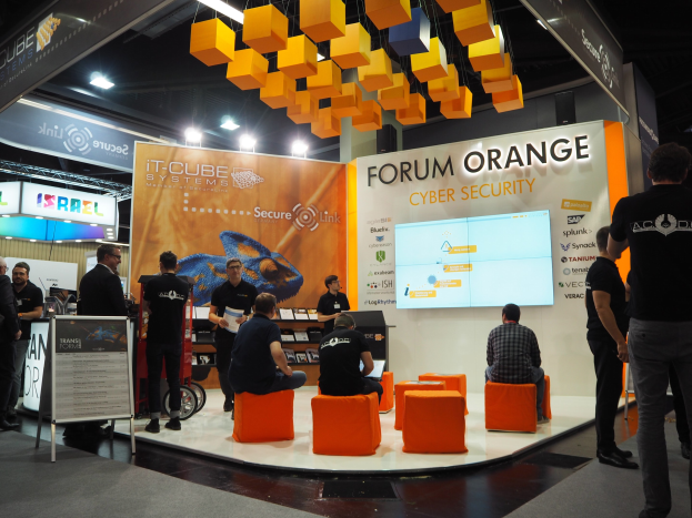 Eine Gruppe von Menschen an der IT-Cube Systems Forum Orange Cyber Security Theke, einige sitzen, andere stehen, umgeben von Anzeigen, Texttafeln, Regalen und Deckenleuchten.