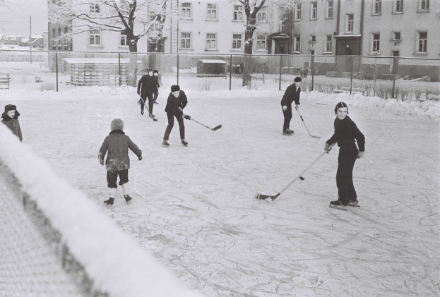 Schwarzes und weißes Bild von Menschen, die Hockey auf einem Eisplatz spielen, der von einem Zaun, Bäumen, Gebäuden und einer Bank umgeben ist.