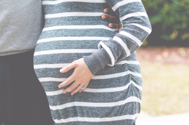 Schwangeres Paar steht Seite an Seite mit den Händen auf dem Bauch, trägt Schwangerschaftskleidung und ist von Bäumen umgeben.