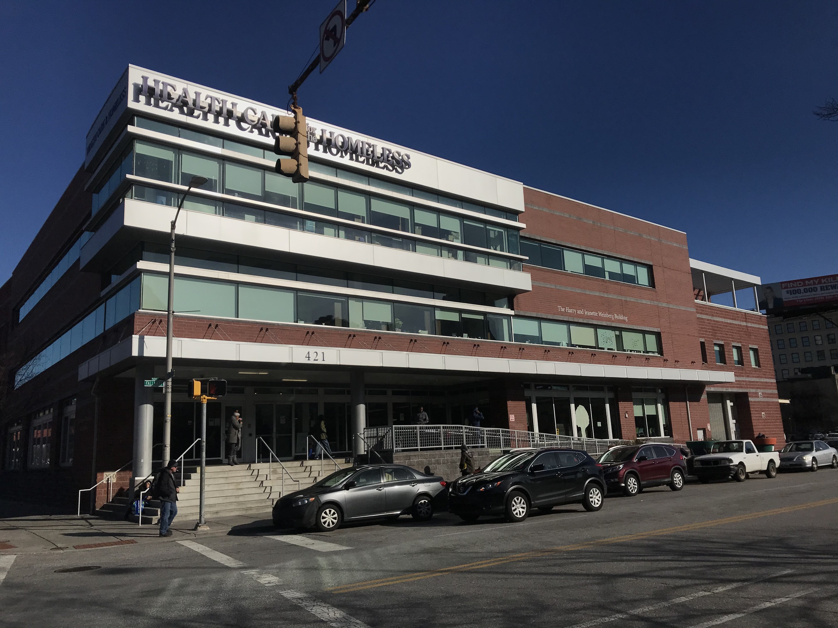 Großes Gebäude als New York City Health Care Obdachlosenzentrum etikettiert mit parkenden Autos, Fußgängern und städtischer Infrastruktur unter einem klaren blauen Himmel.