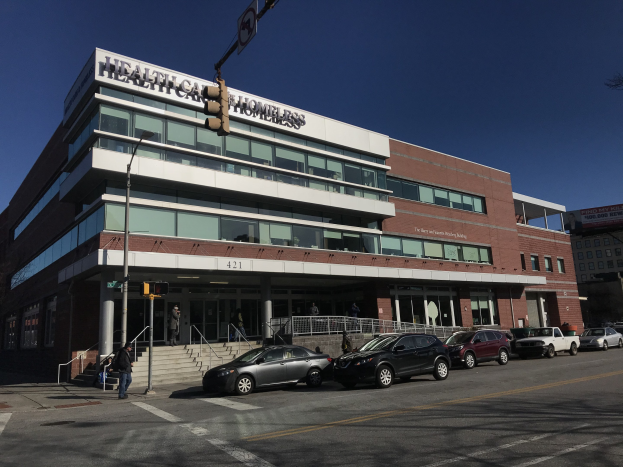 Großes Gebäude als New York City Health Care Obdachlosenzentrum etikettiert mit parkenden Autos, Fußgängern und städtischer Infrastruktur unter einem klaren blauen Himmel.