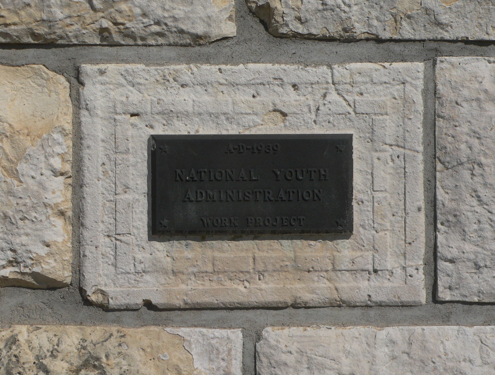 Steinwand mit einem rechteckigen Schild, auf dem in weißer, fetter Schrift "Nationaler Jugenddienst" steht.