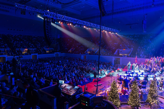 Eine große Menschenmenge in einem geschmückten Auditorium schaut einer Band auf einer festlichen Bühne mit Weihnachtsbäumen, Lautsprechern und beleuchteter Decke zu.