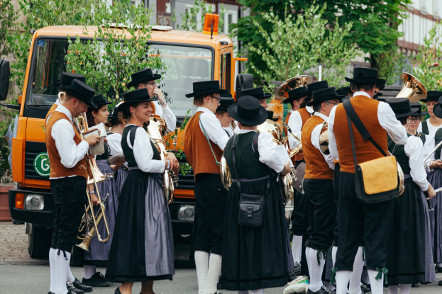 Gruppe von Menschen in traditioneller bayerischer Tracht, die die Straße entlangmarschieren und Blasinstrumente spielen und Taschen tragen, mit Bäumen und Gebäuden im Hintergrund und einem geparkten Fahrzeug.