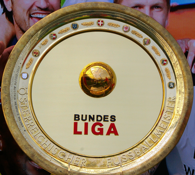 Ein Mann hält einen goldenen Schild mit der Aufschrift "Bundesliga" hoch, mit einem Banner im Hintergrund, auf dem drei Personen zu sehen sind.