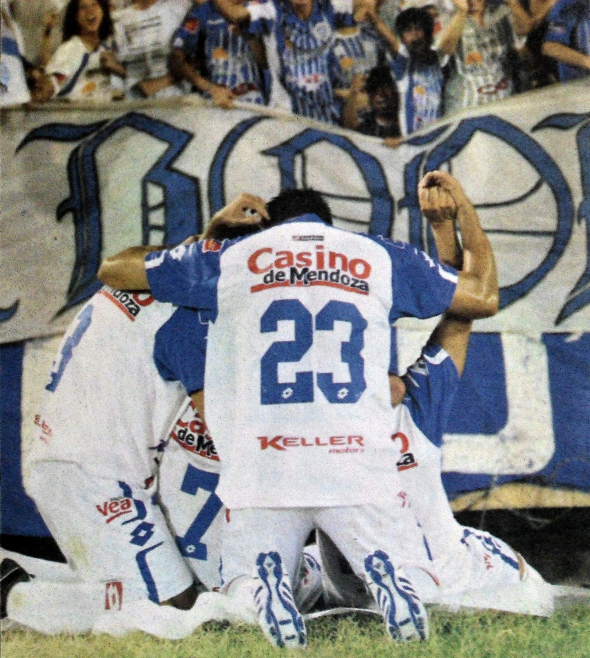 Gruppe von Fussballspielern feiert auf dem Platz vor jubelnder Menge, mit einem Banner mit Text im Hintergrund.