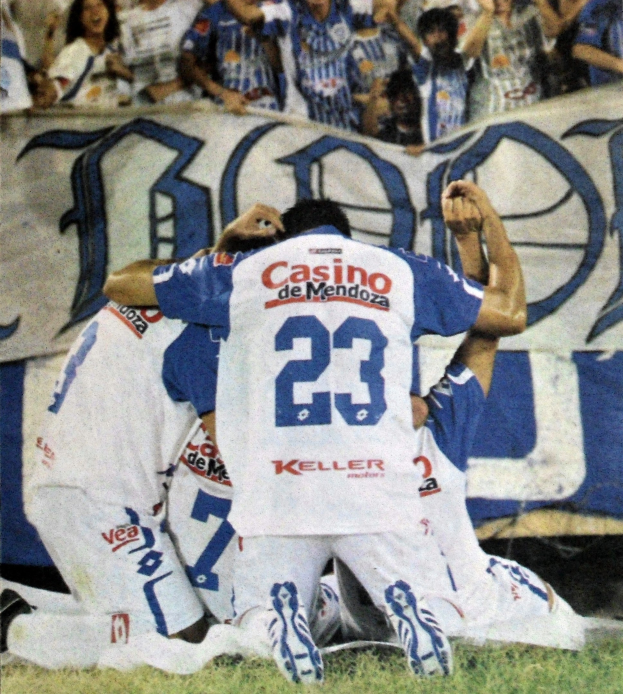 Gruppe von Fussballspielern feiert auf dem Platz vor jubelnder Menge, mit einem Banner mit Text im Hintergrund.