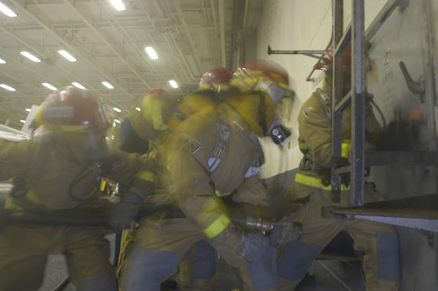 Feuerwehrleute in Helmen und Schutzausrüstung arbeiten an einem Flugzeug, halten verschiedene Gegenstände und haben eine Leiter rechts und Lampen an der Decke im Hintergrund.