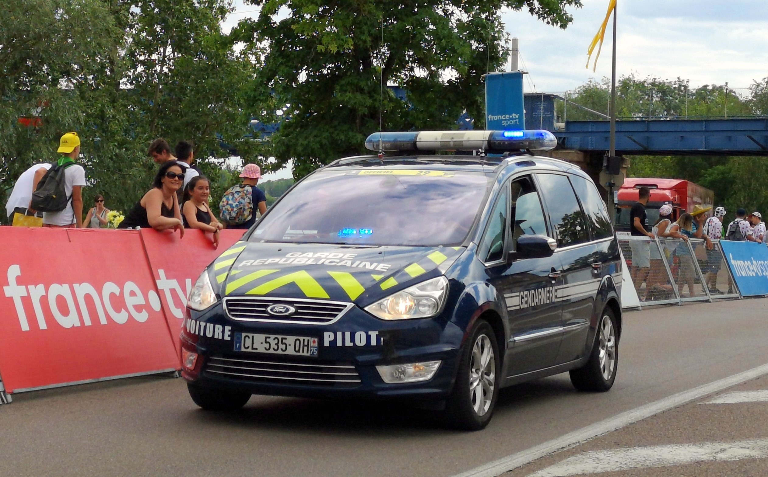 Polizeiauto fährt auf einer Straße neben einer Menge, mit einem Banner links, Gelöndern mit Bannern dahinter, Bäumen, einer Br√∞cke, einer Fahne auf einem Mast und einem bewölktem Himmel im Hintergrund.