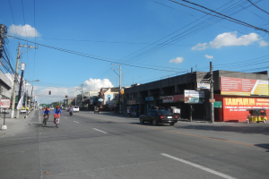 Eine belebte Stra├če mit Menschen auf Fahrr├Ądern und Autos, Strommasten mit Dr├Ąhten, Geb├Ąude mit Namensschildern und ein bew├Âlktes Himmel im Hintergrund.
