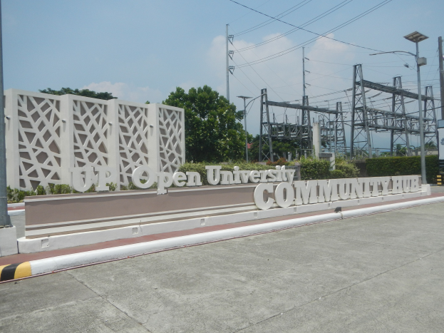 Eingang zum Open University Community Hub mit Text an der Wand, einer Straße, Bäumen, Pflanzen, Laternen, Übertragungsmasten mit Drähten und einem bewölkten Himmel im Hintergrund.