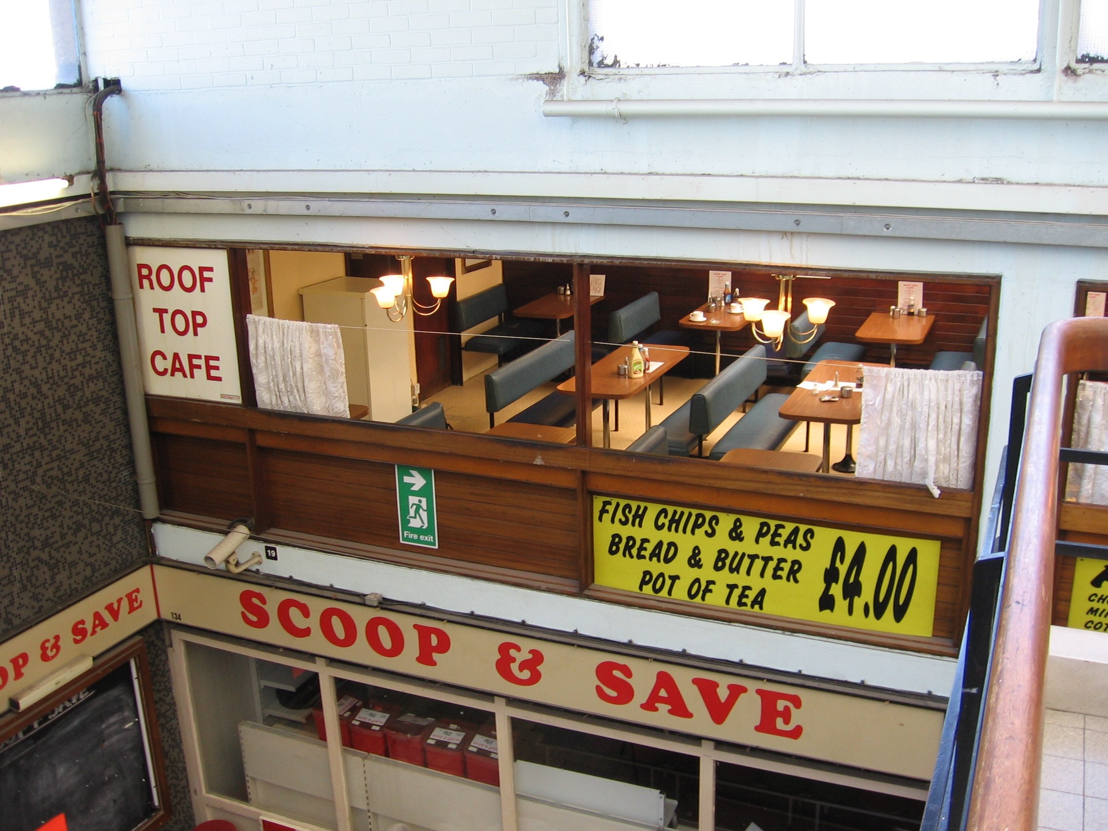 Ein caféartiges Restaurant mit Tischen, Stühlen und beleuchteten Tafeln mit Text, das eine Brüstung auf der rechten Seite und eine Wand im Hintergrund hat und durch ein "Scoop & Save"-Schild gekennzeichnet ist.