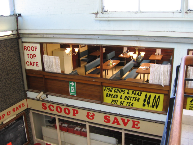 Ein caféartiges Restaurant mit Tischen, Stühlen und beleuchteten Tafeln mit Text, das eine Brüstung auf der rechten Seite und eine Wand im Hintergrund hat und durch ein "Scoop & Save"-Schild gekennzeichnet ist.