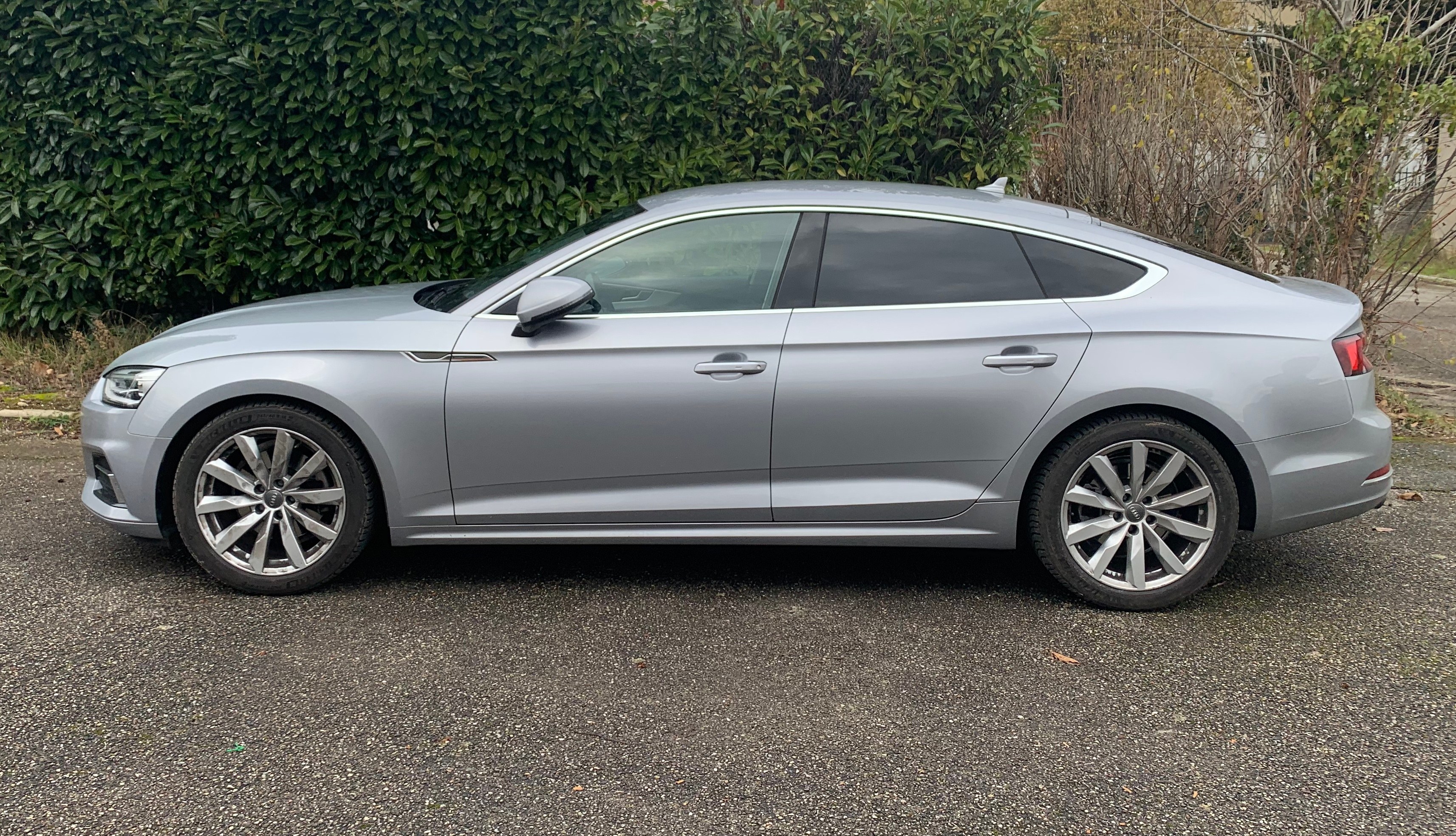 Grauer Audi A5 Sportback 2.0 TDI Quattro Tiptronic S Tronic S Line 5dr Diesel Estate auf einer Straße mit Bäumen und einem Gebäude im Hintergrund.