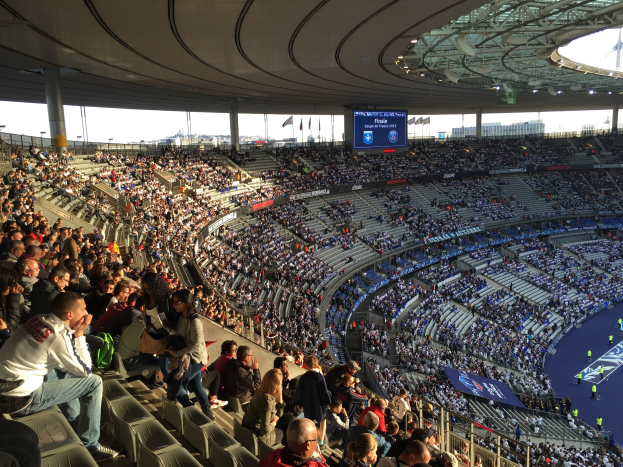 Große Zuschauermenge in einem Stadion bei einem Fussballspiel mit einer Bühne, Fahnen, Stangen, einem Bildschirm und der Allianz Arena in München, Deutschland im Hintergrund.