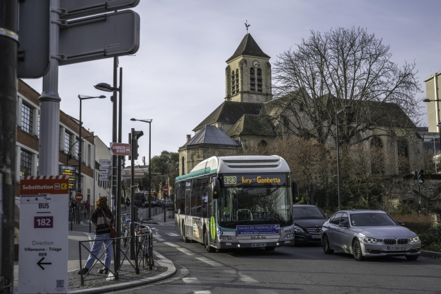 Stadtstraße mit fahrenden Autos, einem Bus, Füülern auf dem Gehweg, abgestellten Fahrrädern, Schildern an Masten und Gebäuden mit Bäumen unter einem klaren blauen Himmel.