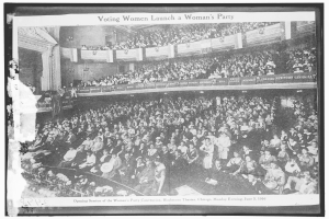 Ein Schwarz-Weiß-Foto einer großen Menschenmenge in einem Gebäude, einige sitzen, einige stehen, Schilder mit Text und eine Wand im Hintergrund. Unten steht der Text "Wahlberechtigte Frauen gründen eine Frauenpartei."