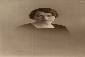 Ein Schwarz-Weiß-Foto einer Frau mit ernstem Gesichtsausdruck, deren Haar zurückgekämmt ist und die eine Kette und ein einfaches Kleid mit hohem Kragen und langen Ärmeln trägt.