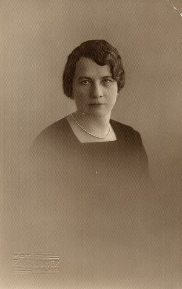 Ein Schwarz-Weiß-Foto einer Frau mit ernstem Gesichtsausdruck, deren Haar zurückgekämmt ist und die eine Kette und ein einfaches Kleid mit hohem Kragen und langen Ärmeln trägt.