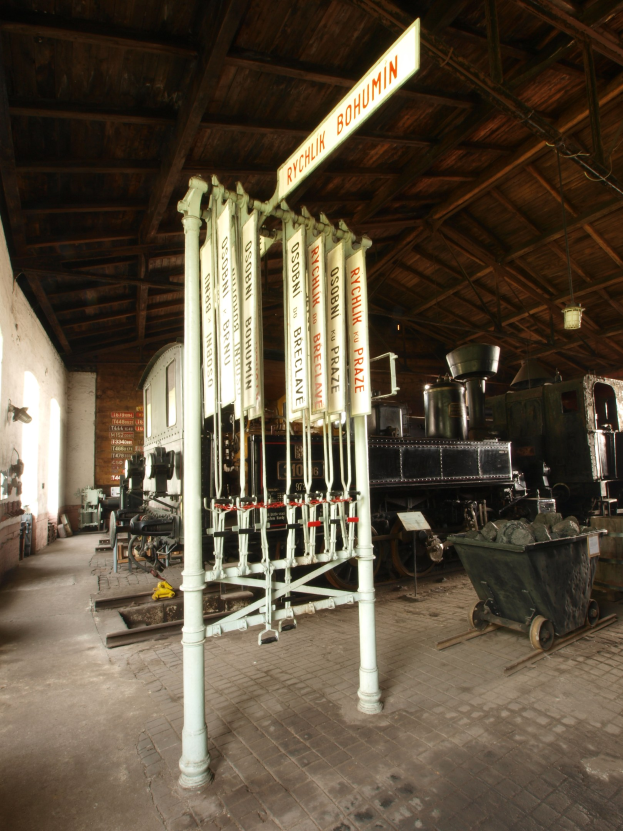 Ein Bahnhof mit einem Zug auf den Schienen, einem Wagen auf dem Boden, einem an einem Pfahl angebrachten Schild, einer Wand auf der linken Seite und einer Decke oben sowie einer zentralen Signalbox, die die Ankunft von Zügen steuert.