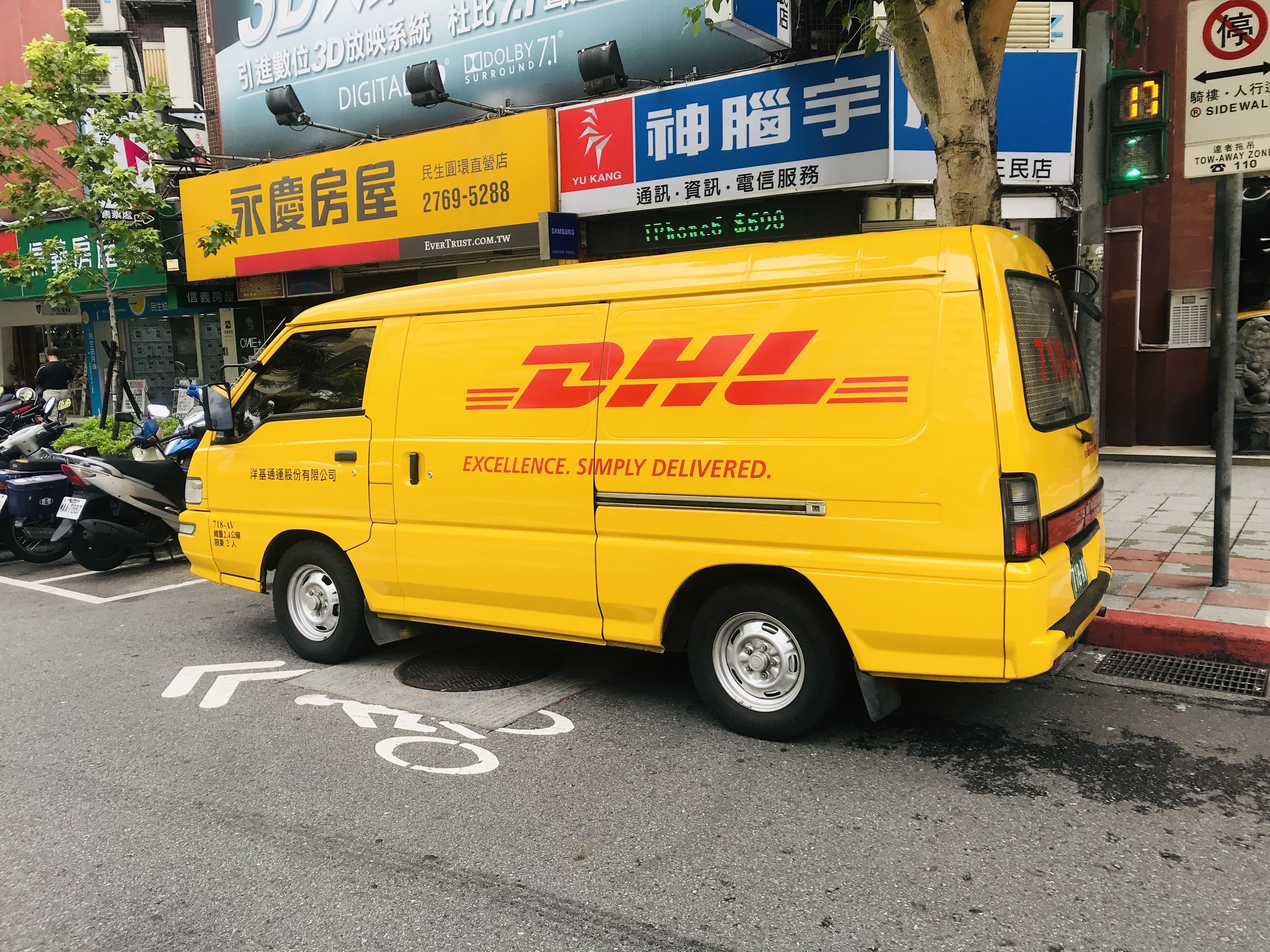Ein gelber DHL-Lieferwagen auf einer Straße geparkt, umgeben von Gebäuden, Bäumen, Schildern, Pfählen, Verkehrsampeln und einem Fußgänger auf dem Gehweg.
