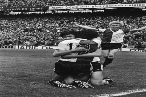 Drei Fußballspieler feiern ein Tor auf dem Boden, mit jubelnder Menge und schwenkenden Fahnen im Hintergrund, dargestellt in Schwarz-Weiß.