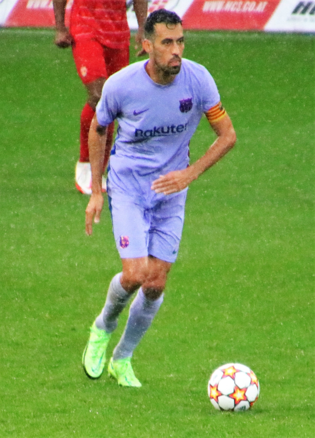 Ein Fußballspieler in Sportbekleidung und -schuhen auf einem Feld mit einer textgefüllten Tafel im Hintergrund.