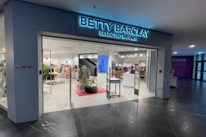 Ein Betty Barclay Selected Brands Geschäft in einem Einkaufszentrum mit Schaufensterpuppen in der neuesten Mode, Kleider an Bügeln, Tischen mit verschiedenen Gegenständen, Schildern, einem Topf mit einer Pflanze, Deckenlampen und einer Tür auf der rechten Seite.