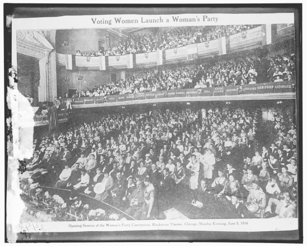 Ein Schwarz-Weiß-Foto einer großen Menschenmenge in einem Gebäude, einige sitzen, einige stehen, Schilder mit Text und eine Wand im Hintergrund. Unten steht der Text "Wahlberechtigte Frauen gründen eine Frauenpartei."