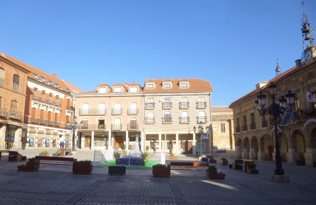 Ein Stadtplatz mit einem zentralen Brunnen, umgeben von Bänken, Topfpflanzen, Straßenlaternen und Lichtern, mit einem Uhrenturm und Gebäuden im Hintergrund unter einem klaren blauen Himmel.