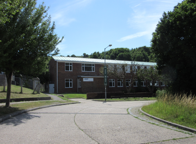 Ein Schulgebäude mit Fenstern, einem Laternenmast, einem Schild, einer Baumgruppe, Gras, einem Zaun und einem bewölkten Himmel, das inmitten eines Parks umgeben von saftigem Grün liegt.